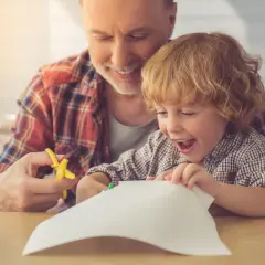 Dziadek pomaga wnuczkowi używać pierwszych nożyczek dla dziecka do wycinania papieru. Radość na twarzy malucha.