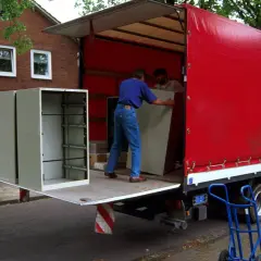 Umzugshelfer laden einen Schrank auf einen roten LKW. Der Schrank steht am Straßenrand, was die Bedeutung von "kerbside" verdeutlicht.