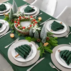 Moderne tischdeko weihnachten: grüne Servietten als Bäume, Tannenzweige, Kerzen und ein Kranz mit Beeren auf einem grünen Tischtuch.