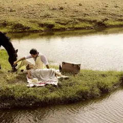 Pareja en un picnic junto a un río, con un caballo pastando. Un momento idílico para disfrutar de rutas en caballo y la naturaleza.
