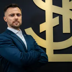Mężczyzna w garniturze, trener ŁKS Łódź, stoi przed złotym logo klubu.