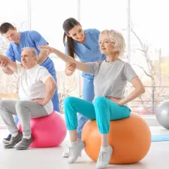 Rehabilitacja dla seniorów: starsza pani i pan ćwiczą z fizjoterapeutami na piłkach gimnastycznych, poprawiając kondycję.