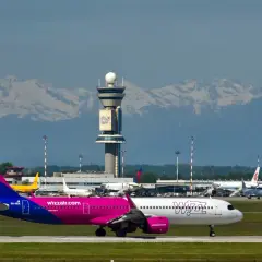 Samolot Wizz Air na płycie lotniska z wieżą kontroli lotów i górami w tle.