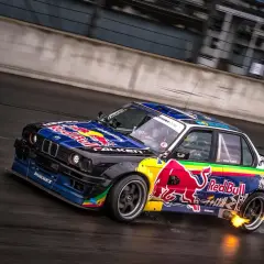 BMW E30 w barwach Red Bull, auto do driftu, z płomieniami z wydechu, na mokrym torze.