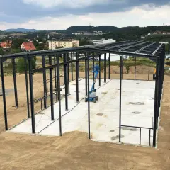 Stalowa konstrukcja hali przemysłowej, gotowa na montaż płyt warstwowych. W tle krajobraz z pagórkami i zabudowaniami.