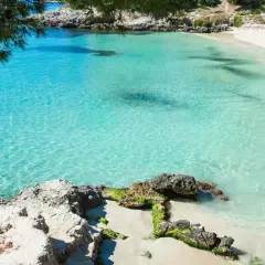 Traumhafter **Palma de Mallorca Strand** mit kristallklarem, türkisfarbenem Wasser und feinem Sand. Felsen und grüne Pinien säumen die Bucht.