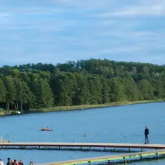 Spokojne jezioro Sierakowo z drewnianymi pomostami, na których stoją ludzie. W oddali widać kajakarzy i zalesiony brzeg.
