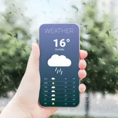 Dłoń trzyma smartfon z aplikacją pogodową pokazującą deszcz i 16 stopni. Za oknem widać krople deszczu.