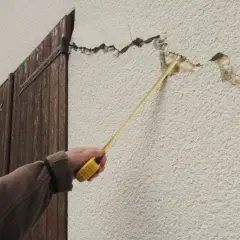 Une main mesure une large fissure sur le mur extérieur d'une maison, près de volets en bois.