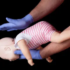 Esercitazione sulla manovra di Heimlich per bambini: mani guantate sostengono un manichino neonato, simulando un intervento di primo soccorso.