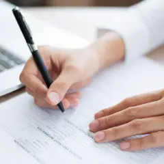 Dłoń pisze długopisem na dokumencie, wypełniając rubryki. Obok leży laptop. To ważne zaświadczenie.