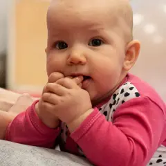 Urocze, 4-miesięczne dziecko w różowej bluzeczce z serduszkami wkłada rączki do buzi, zaciekawione światem.