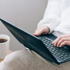 Osoba w białym swetrze pisze na laptopie, obok kubek z kawą i koszyk z włóczką.