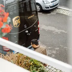 Kurier UPS z paczką zastanawia się, ups gdzie nadać. Obok stoi srebrny samochód.