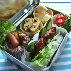 A delicious bento box with a glazed meat skewer, rice, lettuce, egg roll, sausage, beans, corn, tomatoes, and sprouts.
