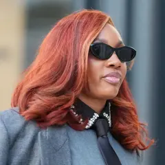 Frau mit feuriger, **cooler Haarfarbe** und stylischer Sonnenbrille trägt einen grauen Blazer mit Krawatte und glitzerndem Kragen.