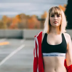 Młoda kobieta w sportowym topie z włókien syntetycznych i czerwonej bluzie zarzuconej na ramiona, na tle jesiennego krajobrazu.