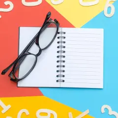 Occhiali neri su un quaderno aperto, circondati da numeri in legno sparsi su sfondi colorati. Un promemoria visivo per imparare l'inglese, magari il numero 66.