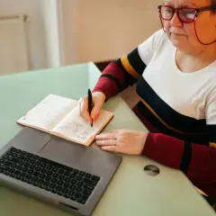 Kobieta w okularach pisze w notatniku obok laptopa.
