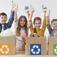 Niños felices participan en un taller de reciclaje, con guantes y pulgares arriba, junto a cajas de cartón con símbolos de reciclaje.