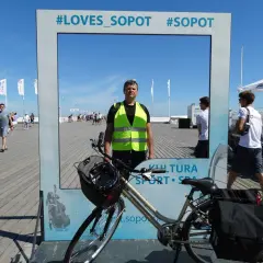 Mężczyzna w kamizelce odblaskowej pozuje przy rowerze na tle ramki z napisem #LOVES_SOPOT. Idealne miejsce na zdjęcie po zwiedzaniu tras rowerowych Sopot.