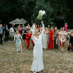 Panna młoda w białej sukni rzuca bukietem, a zgromadzeni goście, w tym panie w kolorowych kreacjach, czekają na jego złapanie. To piękny moment celebrujący tradycje weselne.