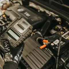 Rękawiczki mechanika pokazują tester akumulatorów, gotowy do sprawdzenia, jak naładować akumulator w samochodzie.