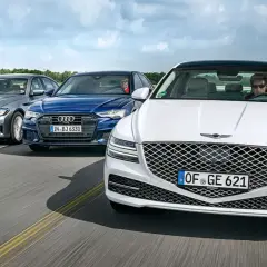Un Genesis blanco lidera un grupo de sedanes de lujo, incluyendo un Audi azul y un BMW gris, en una carretera abierta.