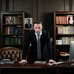 Un homme en costume se tient derrière un bureau, prêt à rendre un jugement contradictoire. Des livres et un drapeau américain ornent la pièce.