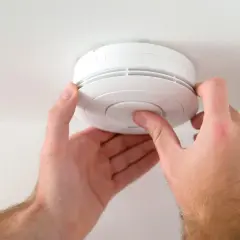 Hände drücken auf den Knopf eines weißen Rauchmelders an der Decke, um den test rauchmelder durchzuführen.