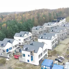 Osiedle domów w budowie na sianowskiej polanie, otoczone jesiennym lasem. Widać rusztowania, materiały budowlane i maszyny.