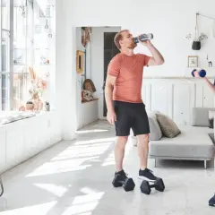 Un couple fait de l'exercice biceps maison avec des haltères bleus dans un salon lumineux.