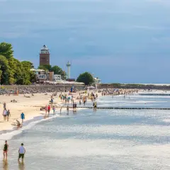 Tłumy ludzi na piaszczystej plaży, latarnia morska w tle. Idealne miejsce na tanie miejscowości nad morzem.