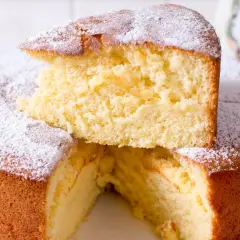 En fluffig vegansk sockerkaka, pudrad med florsocker. En bit är upplagd ovanpå resten av kakan.