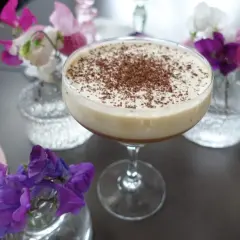 En krämig drink med chokladströssel, perfekt för en stunds avkoppling. Kanske en Kaffe Karlsson? Blommor i rosa och lila ger en vacker inramning.