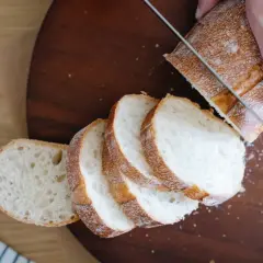 Ręka kroi chleb na desce, widoczne są kromki z białym miękiszem.