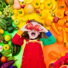Enfant joyeux avec des pommes devant les yeux, entouré de fruits et légumes colorés. Une belle scène pour une activité manuelle semaine du goût.