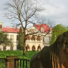 Kamienna rzeźba twarzy przed pałacem w Młoszowej. Budynek z czerwonym dachem i arkadowym gankiem otoczony zielenią.