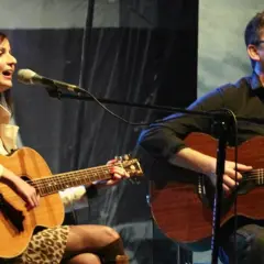 Andrzej Korycki prywatnie, z gitarą i mikrofonem, śpiewa na scenie u boku kobiety grającej na gitarze.