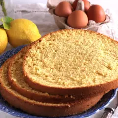 Fette di soffice sponge cake su un piatto blu, con limoni e uova sullo sfondo, pronte per una deliziosa torta.