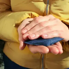 Manos cálidas con un calientamanos reutilizable azul, envuelto en una chaqueta amarilla.