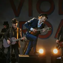 Musicisti sul palco, un uomo canta una canzone d'amore con un mandolino, mentre altri suonano chitarra e basso.