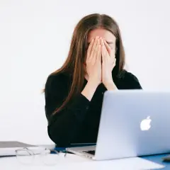 Mujer frustrada cubre su rostro, frente a un portátil. ¿Qué es un falso autónomo? Ejemplos de precariedad laboral.