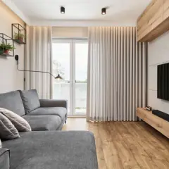 Nowoczesny salon w bloku z szarą kanapą, półkami z książkami i roślinami, telewizorem na tle betonowej ściany i drewnianą szafką.