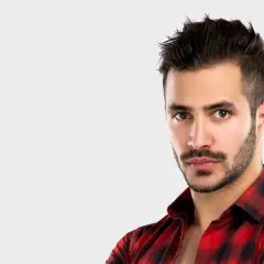 Un homme au regard intense, avec une barbe mouche bien taillée, porte une chemise à carreaux rouges et noirs.