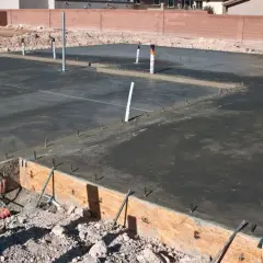 Świeżo wylany beton na płycie fundamentowej, widoczne rury i zbrojenie. Trwa izolacja pozioma fundamentów.