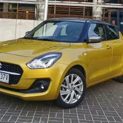 Złoty Suzuki Swift na tle nowoczesnego budynku. Wiele pozytywnych suzuki swift opinie potwierdza jego miejski charakter.