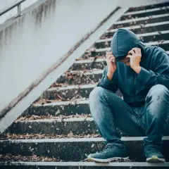 Młoda osoba w kapturze siedzi na schodach, pogrążona w rozpaczy. Obraz symbolizuje mechanizmy uzależnienia od narkotyków i walkę z nimi.