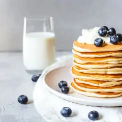 Ein Stapel goldbrauner Protein Pfannkuchen, garniert mit Sahne und Blaubeeren, daneben ein Glas Milch.