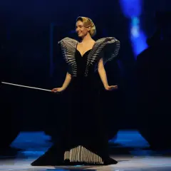 Matilda De Angelis in einem dramatischen schwarzen Kleid mit auffälligen Schulterdetails und einem Dirigentenstab.